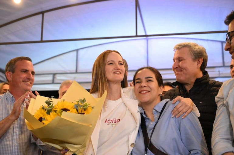Soledad Núñez con blusa blanca y ramo de flores amarillas, sonríe abrazada a Johanna Ortega, con Rafael Filizzola y Stephan Rasmussen detrás.