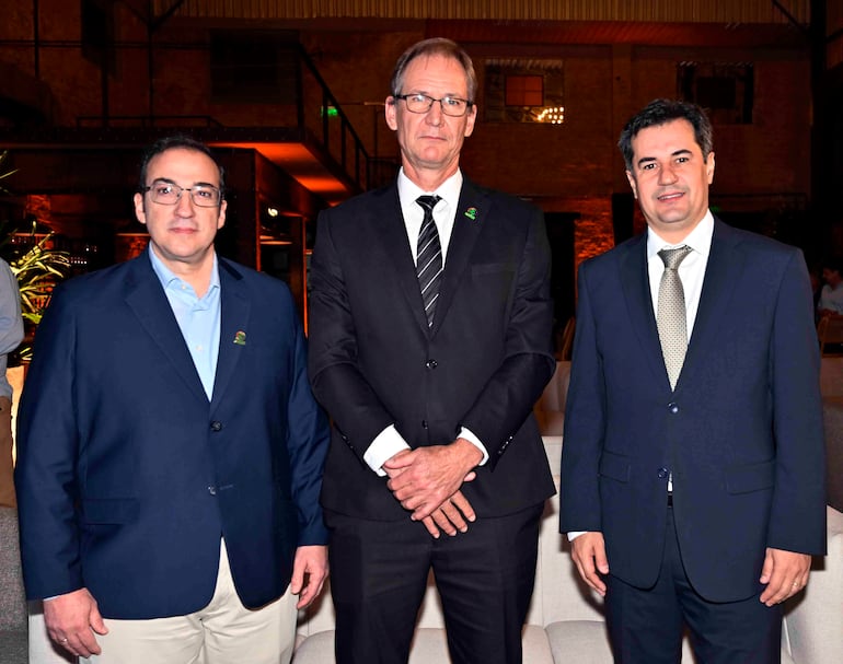 Tres hombres en un salón elegante: uno con blazer azul, otro con traje negro y corbata a rayas, y otro en traje azul oscuro.