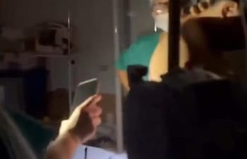 Tres personas en quirófano; una con camisa verde asiste al paciente, otra sostiene un celular para iluminar.
