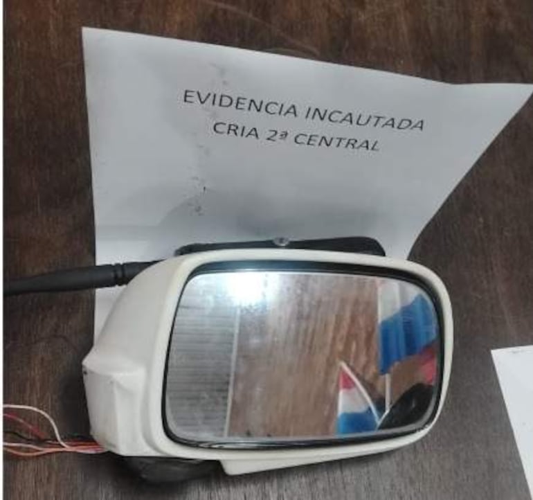 Espejo lateral de auto sobre superficie de madera junto a un documento que dice 'EVIDENCIA INCAUTADA CRIA 2ª CENTRAL'.