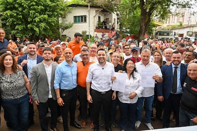 Camilo Pérez sonríe mientras sostiene papeles, rodeado de personas con camisetas naranjas y azul en un ambiente al aire libre.