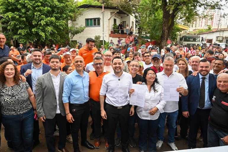 Camilo Pérez sonríe mientras sostiene papeles, rodeado de personas con camisetas naranjas y azul en un ambiente al aire libre.