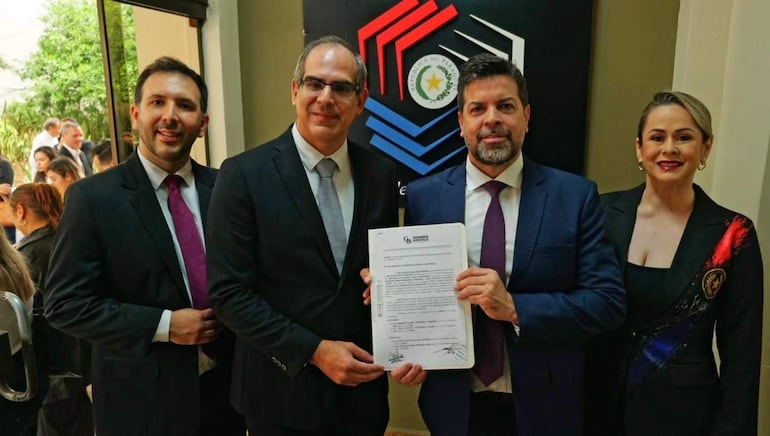 Gerardo Bobadilla sonríe en traje negro, mientras César Escobar serio junto a José Antonio Valenzuela que sostiene un documento, y Zully Villalba sonríe.