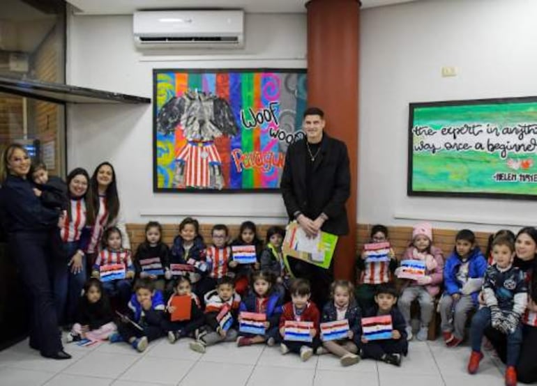 Grupo de niños con camisetas rojas y blancas sostiene banderas de Paraguay; adultos interactuando en un ambiente colorido.