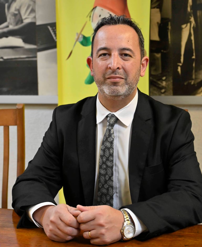 Hombre con traje negro y corbata oscura, sentado en mesa de madera, en ambiente formal y privado.
