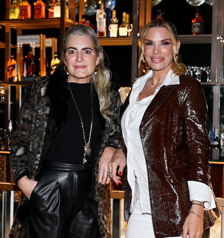 Mujer con abrigo de leopardo y otra con blazer marrón sonríen en un bar, rodeadas de botellas y copas.