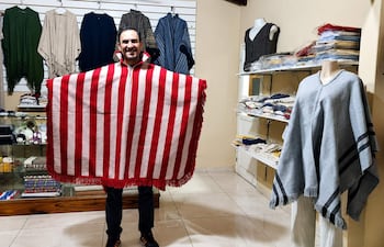 Marco Ramírez sonríe sosteniendo un poncho albirrojo en una tienda, con estantes llenos de ropa detrás.