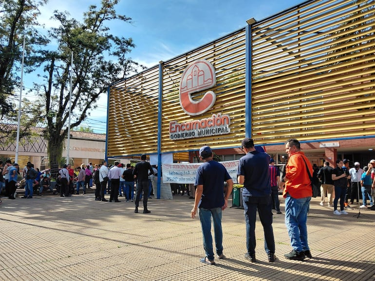 Grupo de hombres espera en fila frente al Gobierno Municipal de Encarnación, con camisetas oscuras y vestimenta informal, en un ambiente diurno.