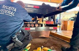 Tres agentes de aduanas revisan un camión, rodeados de cajas con plástico amarillo y verde en un puesto de control.
