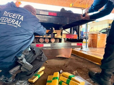 Tres agentes de aduanas revisan un camión, rodeados de cajas con plástico amarillo y verde en un puesto de control.