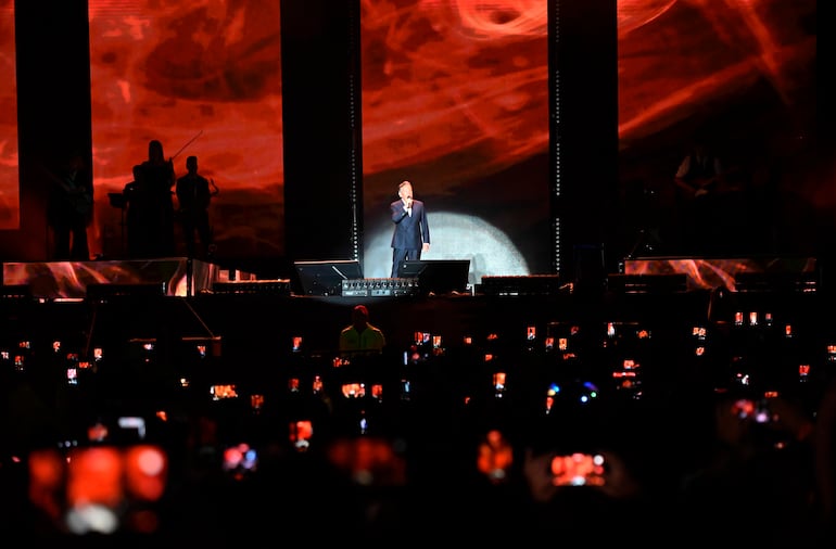 Montaner con traje oscuro en el escenario iluminado, rodeado de músicos mientras la multitud graba con sus teléfonos.