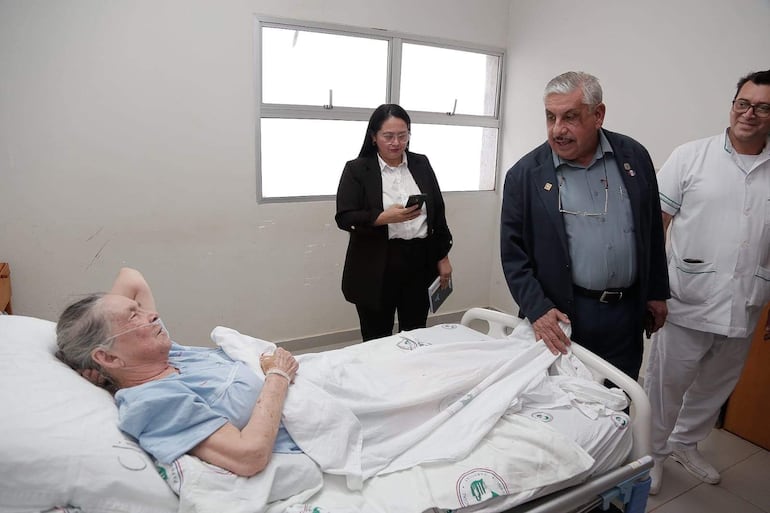 Isaías Ricardo Fretes, en traje oscuro, conversa con mujer mayor en cama con mascarilla de oxígeno, mientras un médico observa.