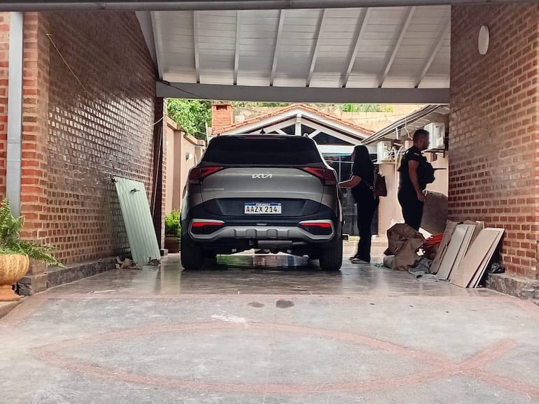 Dos personas conversan junto a un SUV en un garaje, mientras el entorno muestra escombros y ladrillos.