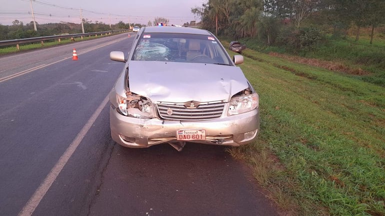 Sedán plateado dañado con capó doblado en un camino rural rodeado de vegetación.