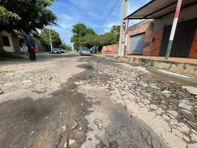 Hombre de pie en abrigo oscuro observa la calle deteriorada con charcos de agua y árboles al fondo.