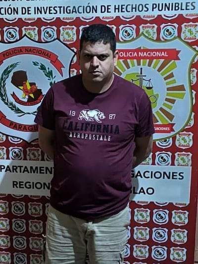 Hombre de pie frente al escudo de la Policía Nacional, vestido con camiseta burdeos y pantalones cortos claros, muestra expresión seria.