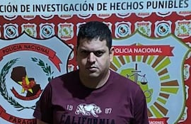 Hombre de pie frente al escudo de la Policía Nacional, vestido con camiseta burdeos y pantalones cortos claros, muestra expresión seria.