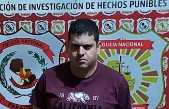 Hombre de pie frente al escudo de la Policía Nacional, vestido con camiseta burdeos y pantalones cortos claros, muestra expresión seria.