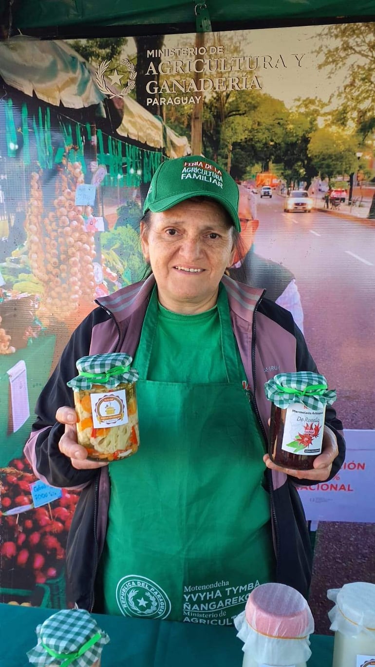 Mujer con gorra del Ministerio de Agricultura sonríe mientras sostiene frascos de mermelada en un evento al aire libre.