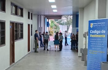 Tres adultos conversan cerca de una mesa mientras una estudiante en uniforme habla con un guardia en un pasillo iluminado.