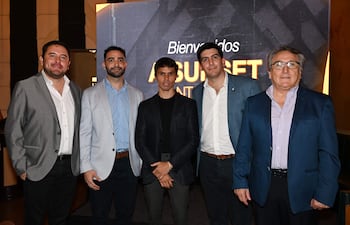 Cinco hombres, uno en camiseta negra y otros en trajes, sonríen en un evento con un cartel de bienvenida al fondo.