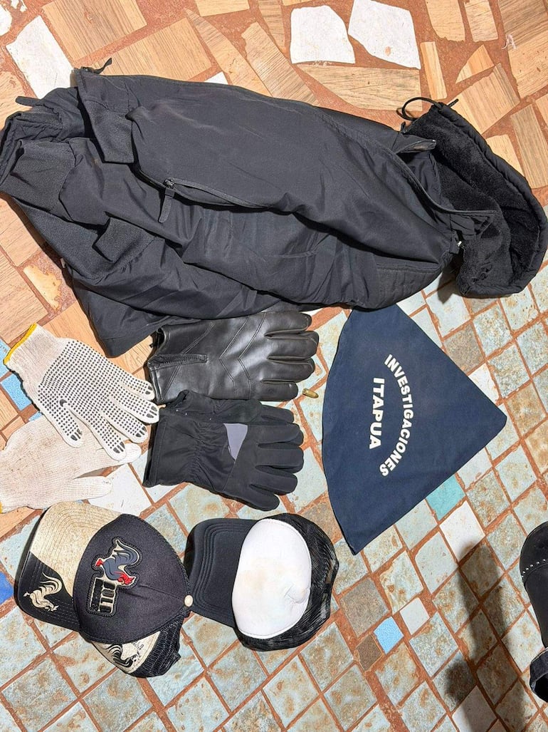 Objetos de investigación sobre asalto: chaqueta negra, guantes y gorras sobre baldosa, con pañuelo que dice 'Investigaciones Itapúa'.