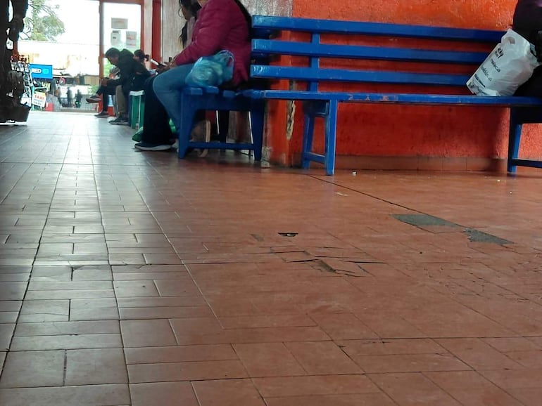 Personas sentadas en bancos azules en un terminal con piso de baldosas y paredes naranjas, una con mochila revisando su teléfono.