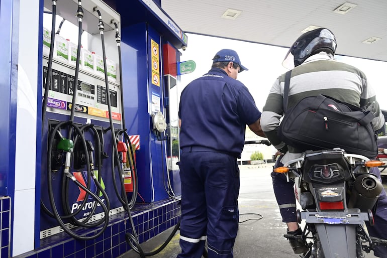 Empleado de Petropar en uniforme azul atiende a cliente con casco negro y camiseta de rayas en estación de servicios al aire libre.