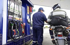 Empleado de Petropar en uniforme azul atiende a cliente con casco negro y camiseta de rayas en estación de servicios al aire libre.