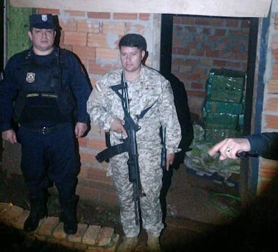Hombre con uniforme policial y otro militar sosteniendo un fusil, observando bultos sospechosos en un entorno oscuro.
