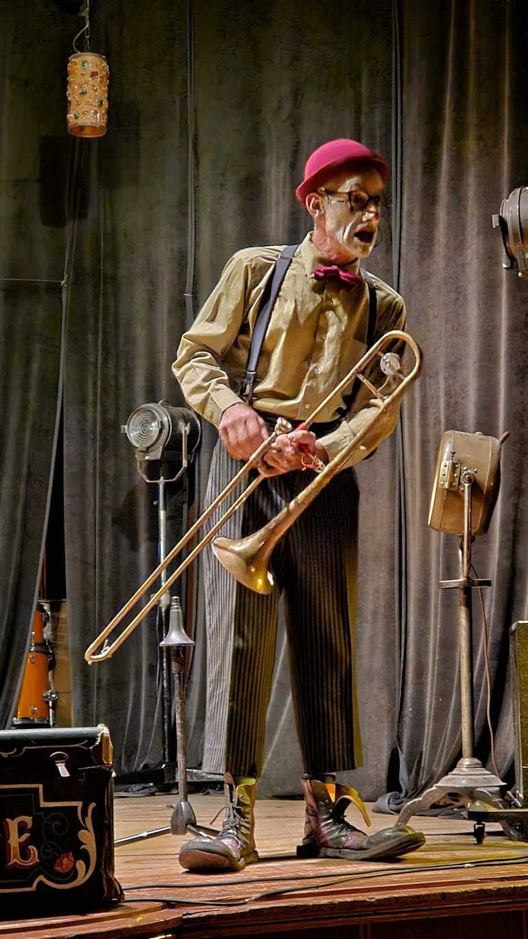 Hombre mayor con sombrero rosa y camisa amarilla toca el trombón, mostrando concentración en un escenario iluminado.