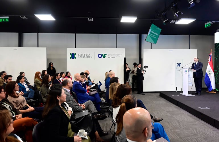 Óscar Lovera en un podio con traje formal, mientras el público escucha atentamente en un auditorio lleno.