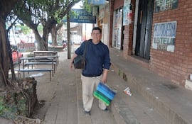 Miguel Giménez, lustrabotas, viste suéter oscuro y pantalones claros, sosteniendo una bolsa mientras camina por la acera urbana.