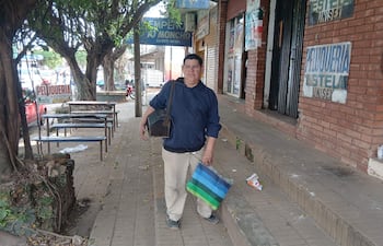 Miguel Giménez, lustrabotas, viste suéter oscuro y pantalones claros, sosteniendo una bolsa mientras camina por la acera urbana.