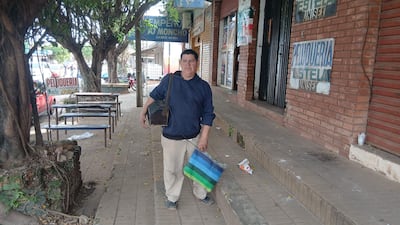 Miguel Giménez, lustrabotas, viste suéter oscuro y pantalones claros, sosteniendo una bolsa mientras camina por la acera urbana.