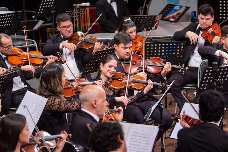 Músicos en trajes formales tocan violines, con una mujer sonriendo en primer plano en un ambiente teatral.