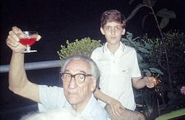 Hombre mayor con gafas sostiene un vaso de cóctel, mientras un niño observa con un petardo en una reunión familiar al aire libre.