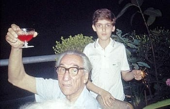 Hombre mayor con gafas sostiene un vaso de cóctel, mientras un niño observa con un petardo en una reunión familiar al aire libre.