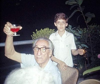Hombre mayor con gafas sostiene un vaso de cóctel, mientras un niño observa con un petardo en una reunión familiar al aire libre.