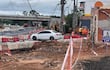 Tres trabajadores en uniforme naranja y casco realizan tareas en un sitio de construcción con un auto blanco estacionado.