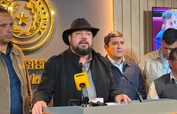 Colym Soroka con gorra negra habla con micrófono, rodeado de hombres en chaquetas oscuras, en un ambiente formal de prensa.