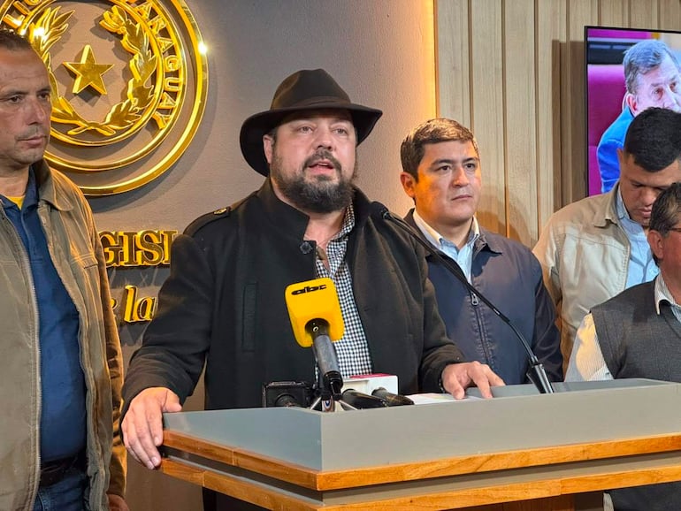 Colym Soroka con gorra negra habla con micrófono, rodeado de hombres en chaquetas oscuras, en un ambiente formal de prensa.