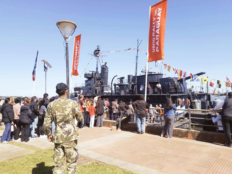 Hombre en uniforme militar observa a la multitud en evento festivo frente al patrullero 'Capitán Cabral'.
