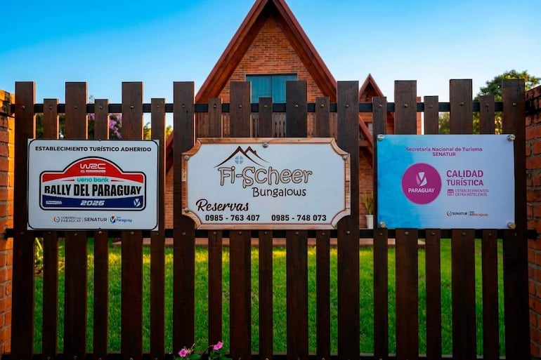 Valla de madera con carteles de Rally del Paraguay 2026 y Fi-Scheer Bungalows, fondo de casa rústica y cielo azul.