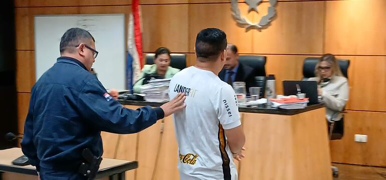 Hombre con camiseta blanca 'LANDERFIT' es sostenido por un policía, mientras cinco personas profesionalmente vestidas observan.