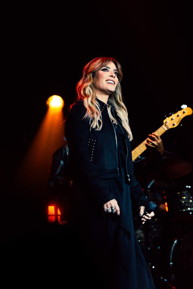 Kany García sonríe mientras sostiene un micrófono, luciendo elegante en el escenario rodeada de luces y músicos.