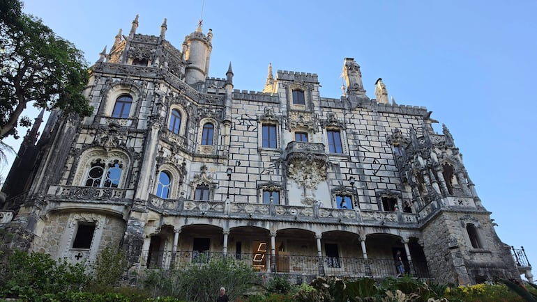 Edificio neogótico con fachada decorada y torres, rodeado de vegetación bajo un cielo despejado.