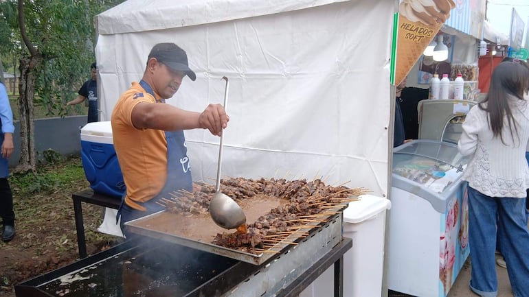 Hombre con camiseta naranja y delantal azul asando carne, en un evento festivo con interacción social y ambiente culinario.