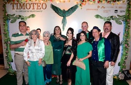 Ocho personas sonríen, posando en un evento, con vestidos coloridos y un fondo decorado relacionado con 'Timoteo'.