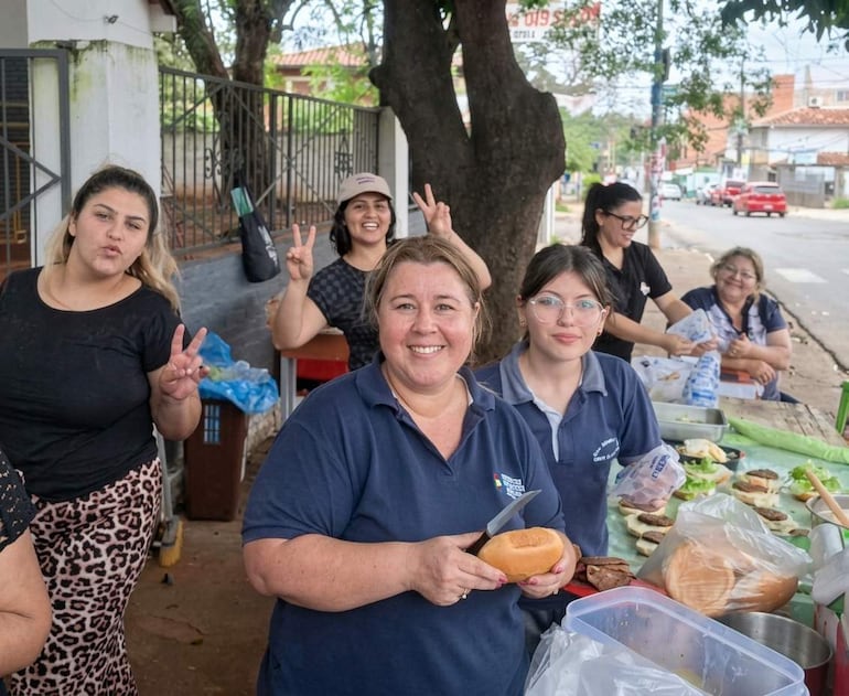 Seis mujeres sonríen en un puesto de comida al aire libre, una sostiene un pan y otra hace el gesto de paz con la mano.